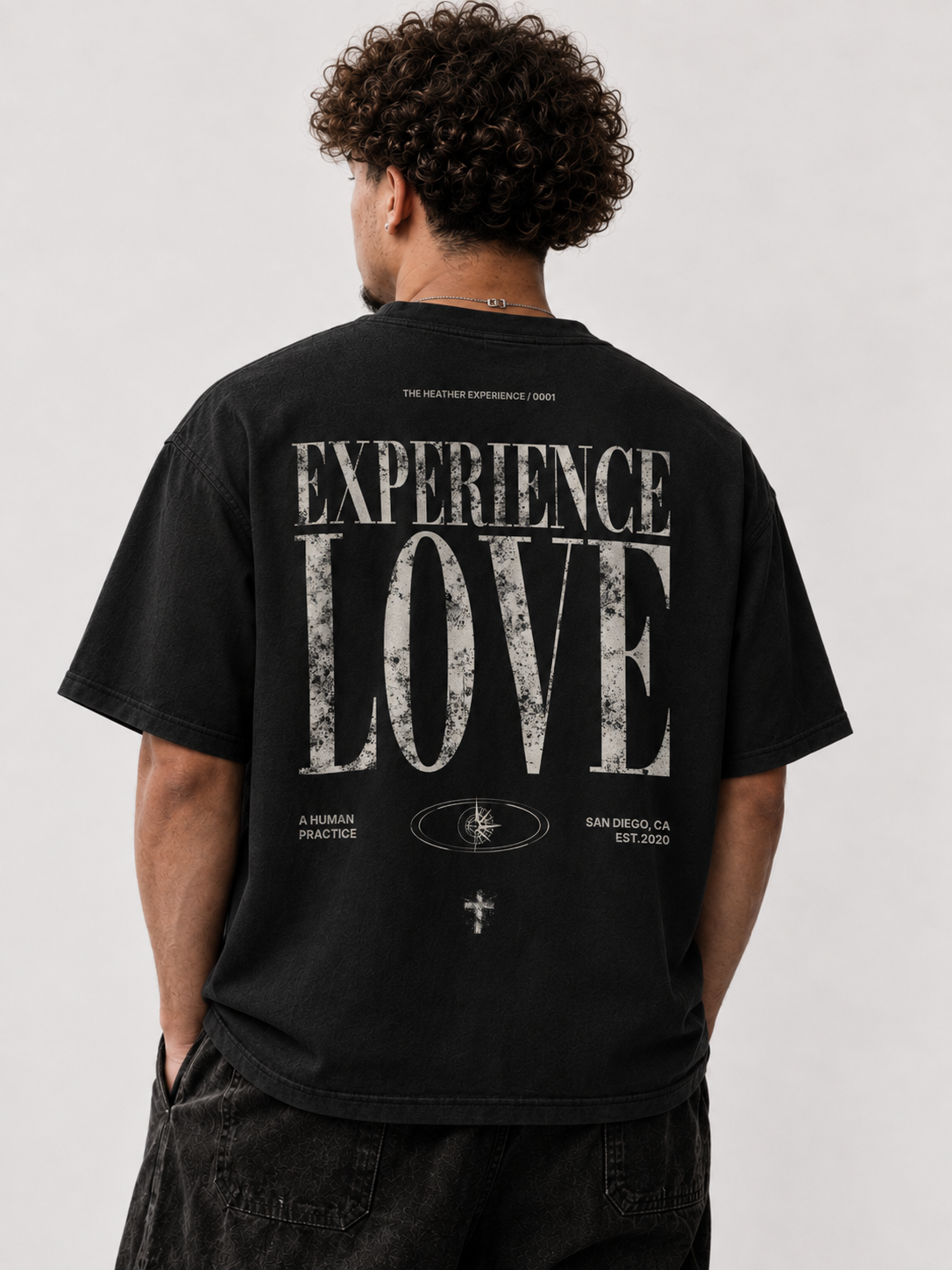 Back of a black EXPERIENCE LOVE T-shirt worn by a model, oversized distressed serif type reading THE HEATHER EXPERIENCE, EXPERIENCE LOVE, A HUMAN PRACTICE, an eye/compass emblem, and SAN DIEGO, CA EST. 2020.