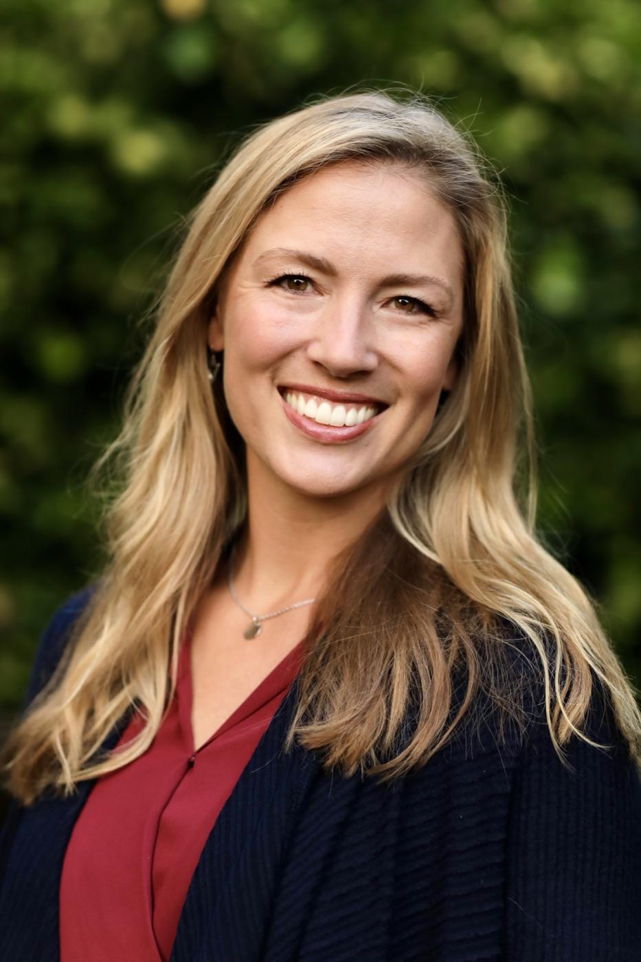 Allie Chapman smiling outdoors in a navy cardigan and burgundy blouse, with green foliage behind her.