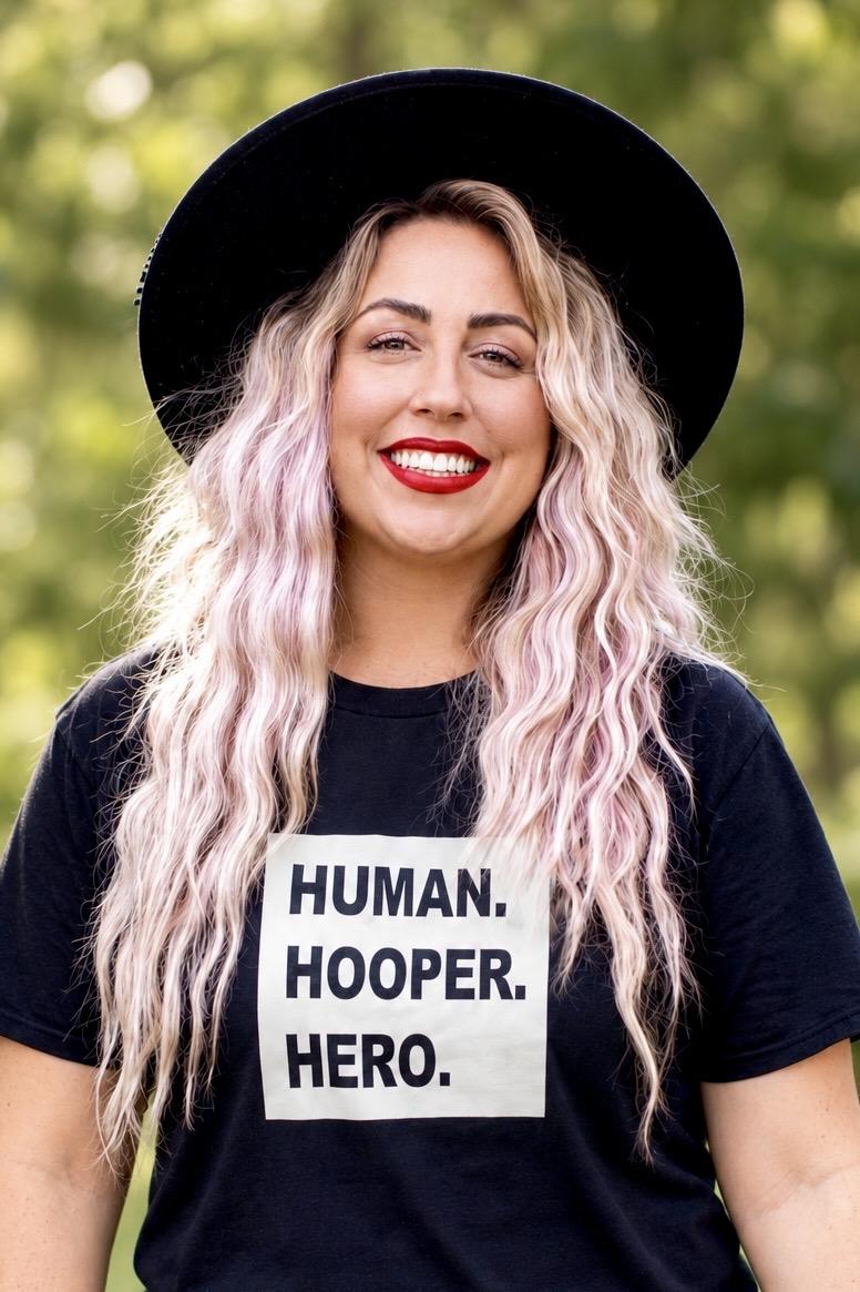 Heather Cleaves smiling outdoors in a black wide-brim hat and a Human. Hooper. Hero. T-shirt.