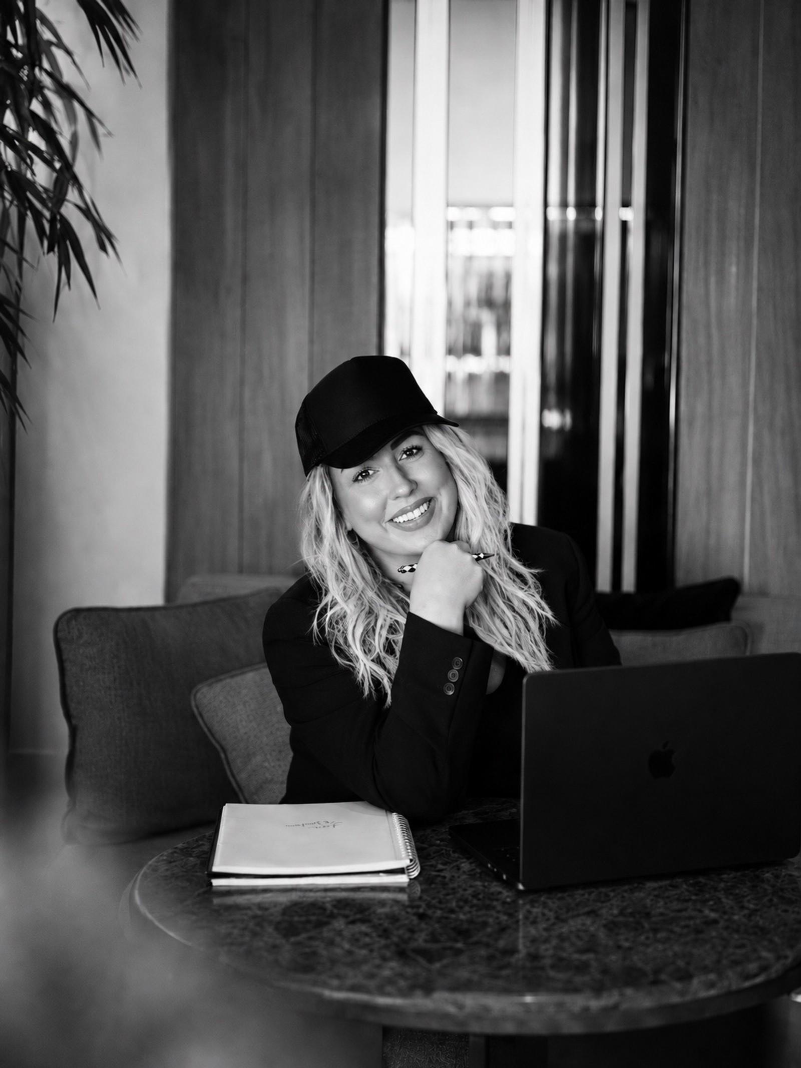 Black-and-white portrait of Heather in a cap and blazer, seated at a table with a laptop and notebook, smiling at the camera.