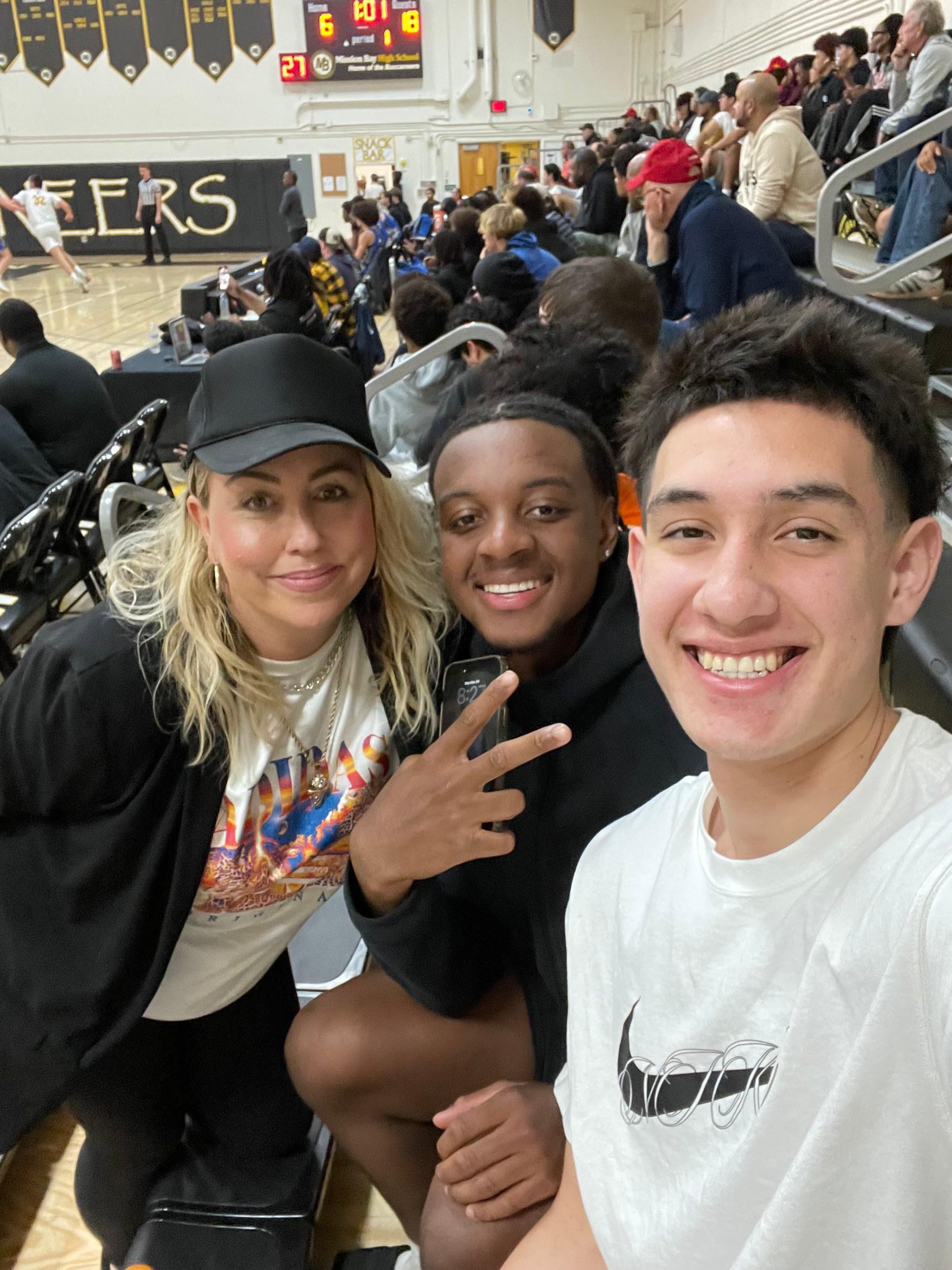Heather courtside with two athletes in a packed gym, smiling together during a game.