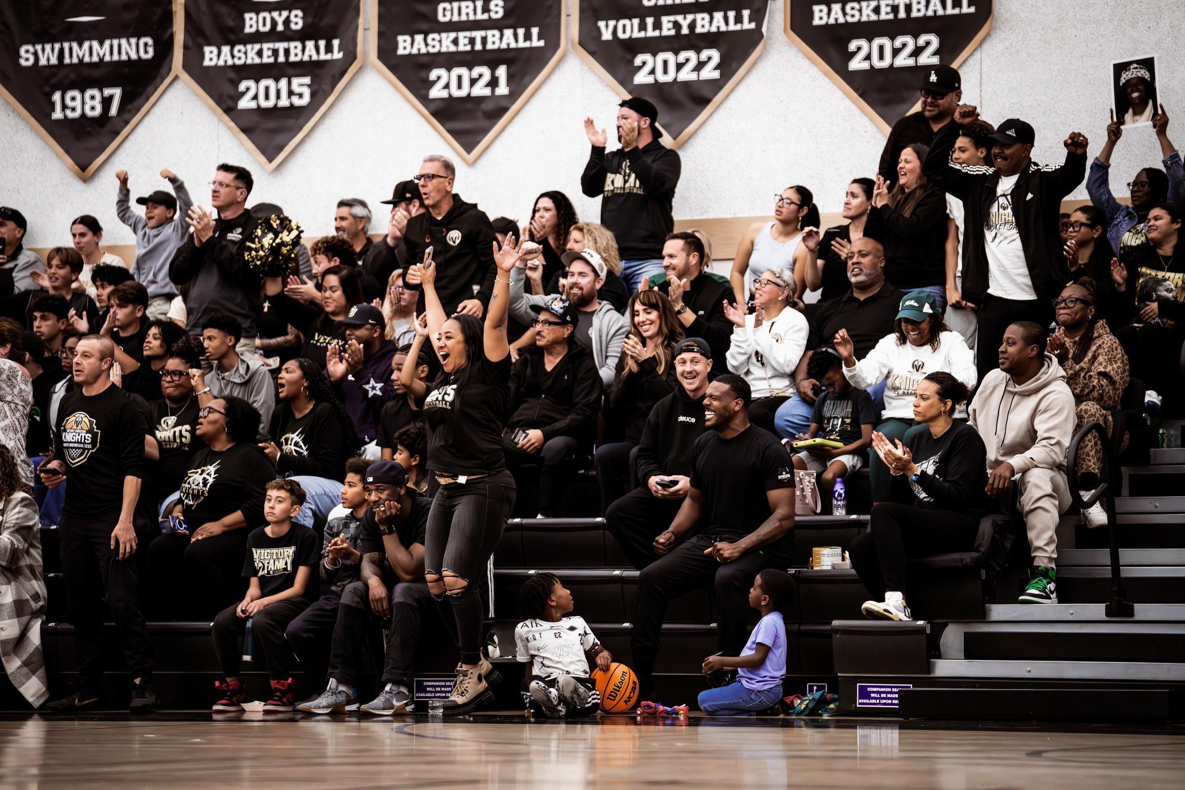 A packed gym of parents and family on their feet cheering, arms raised, championship banners on the wall behind them.